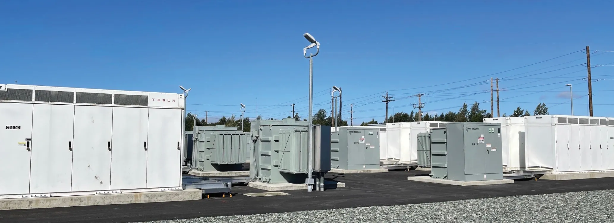 Battery storage units in a facility under a clear blue sky, with power lines and trees in the background.