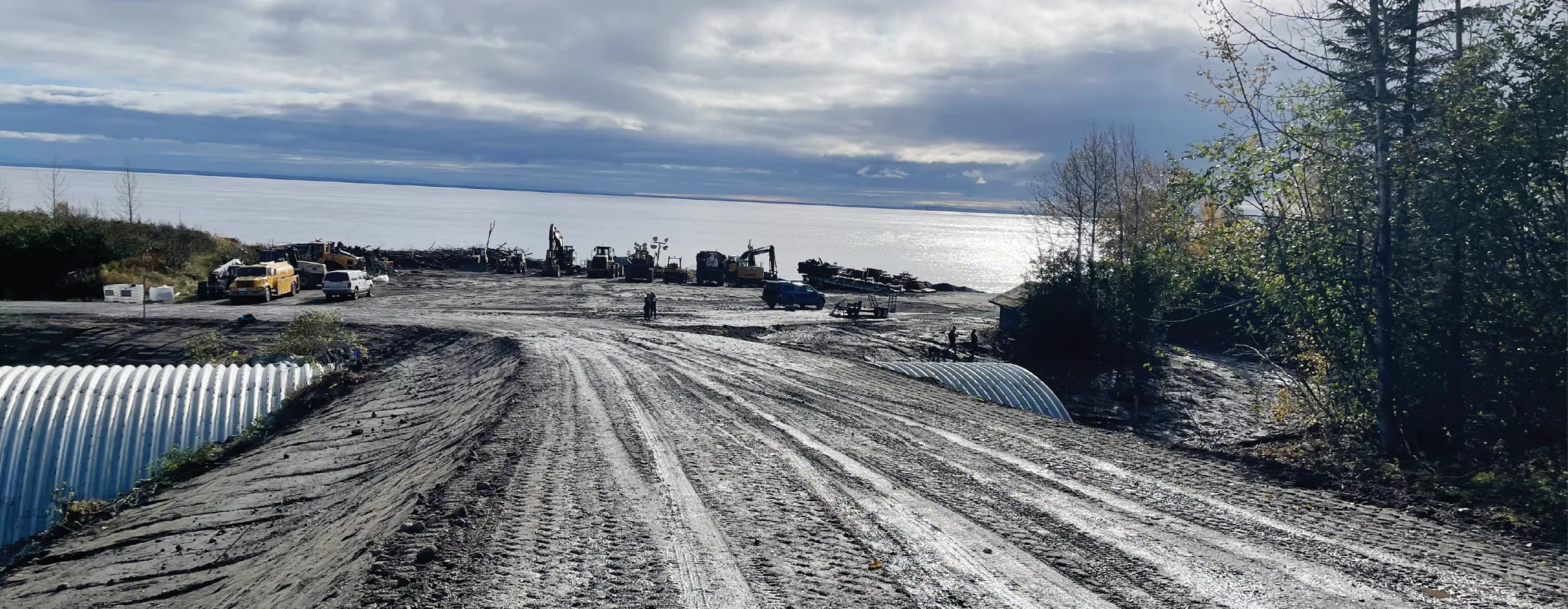 Gravel road leading to a water body with construction equipment and vehicles in the background under a cloudy sky.