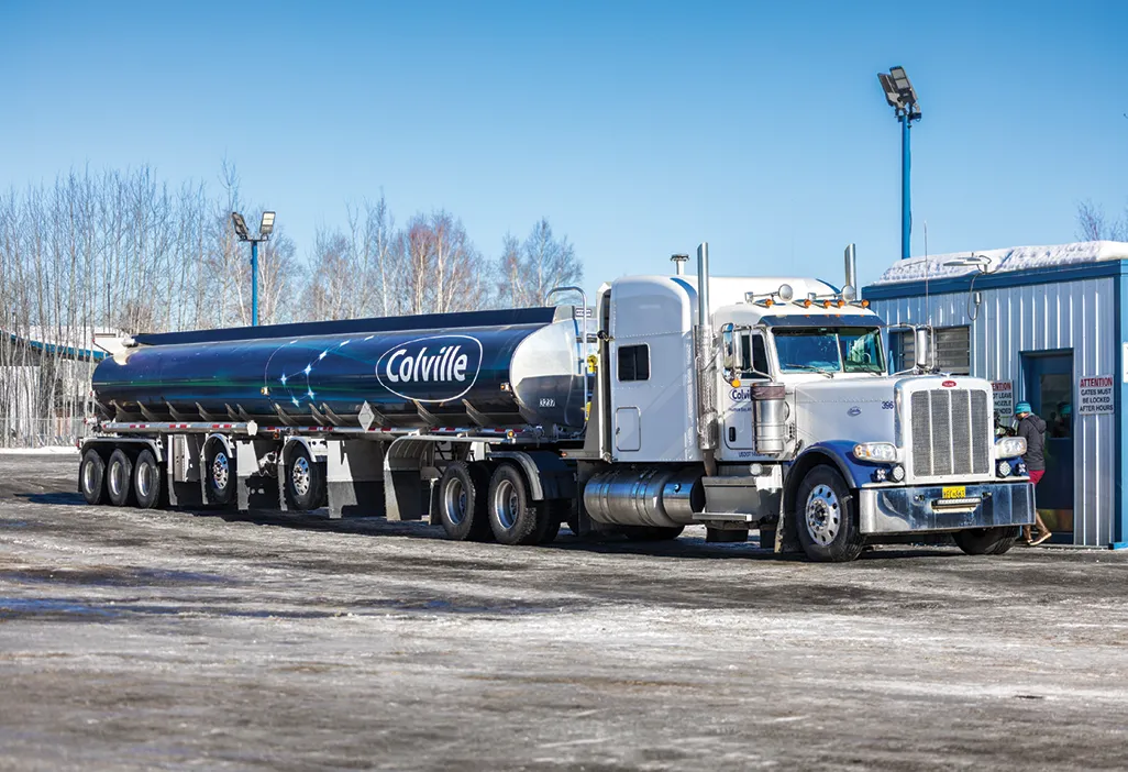 A large white truck with a black tanker trailer labeled "Colville" parked near a building in a snowy area.