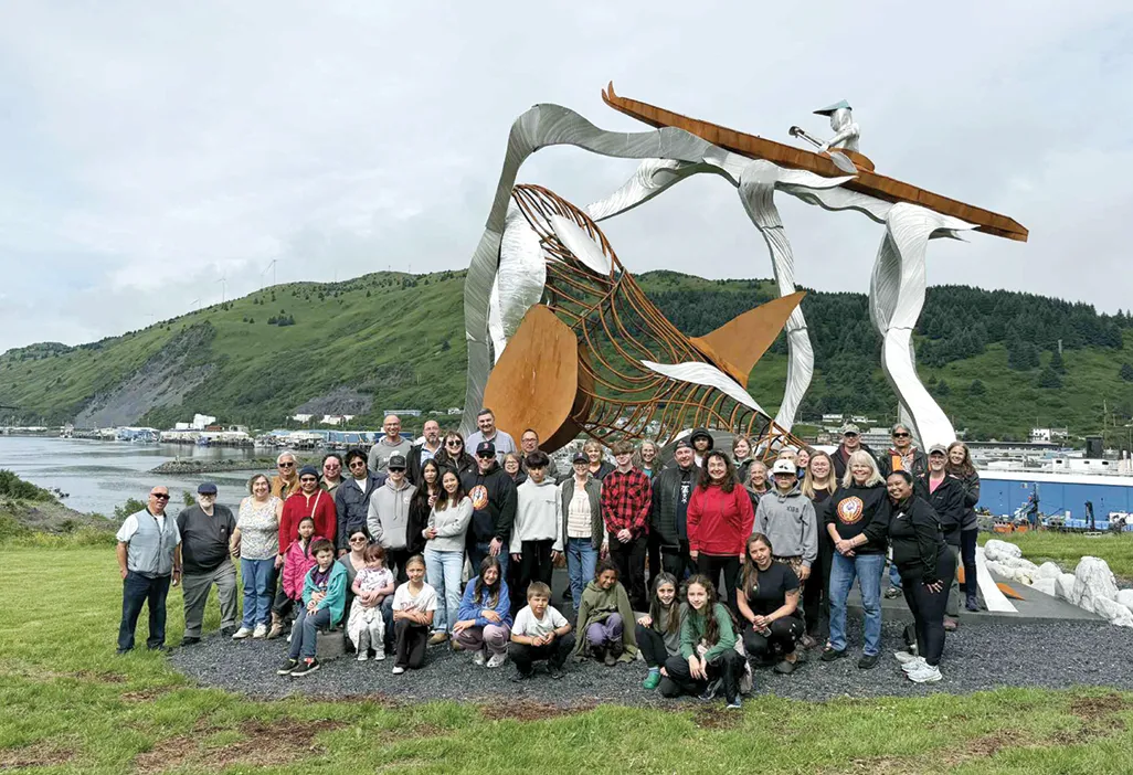 Group of people posing in front of a large, abstract fish sculpture with a mountainous landscape in the background.