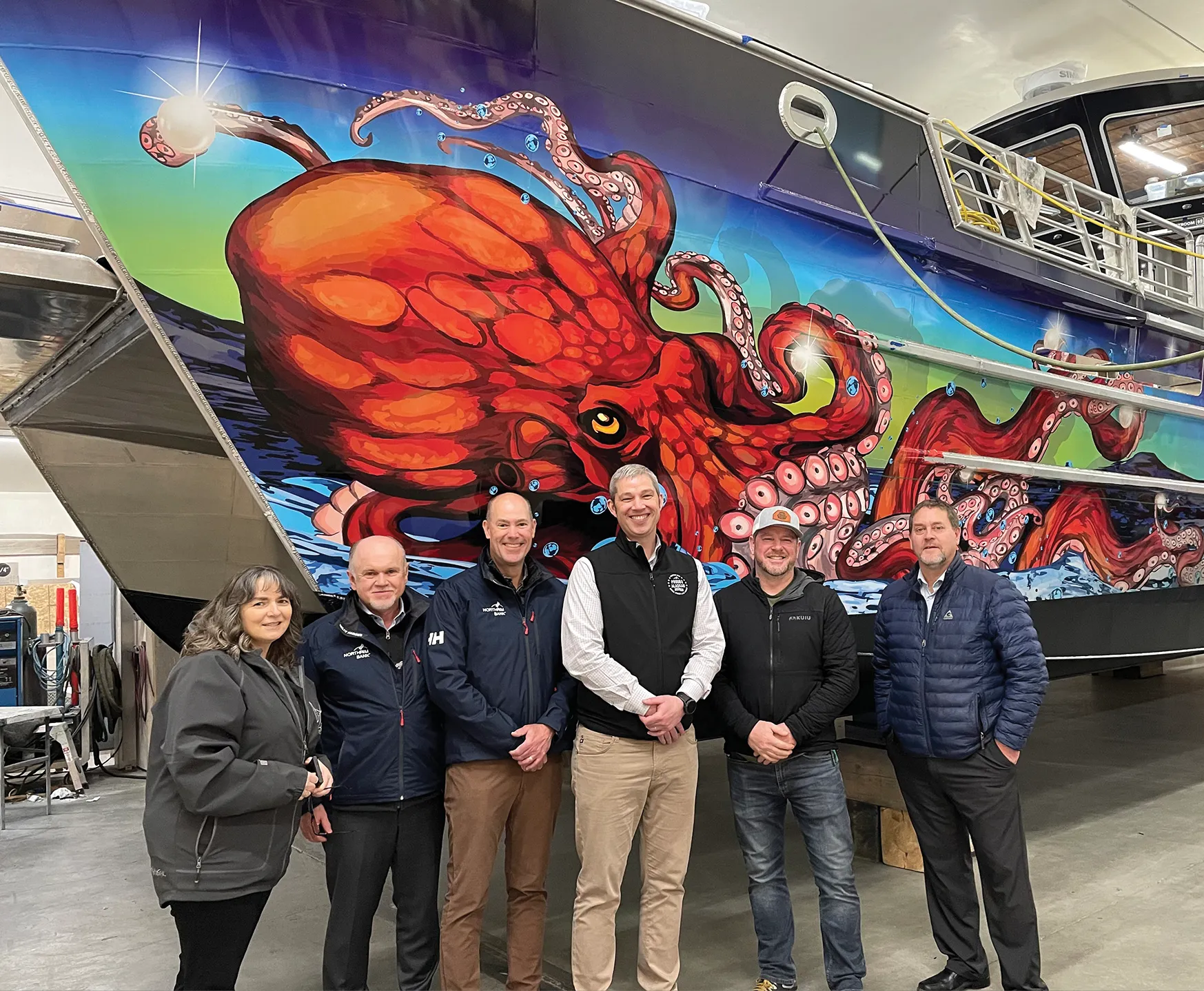 Group of six people posing in front of a colorful octopus mural on a boat.