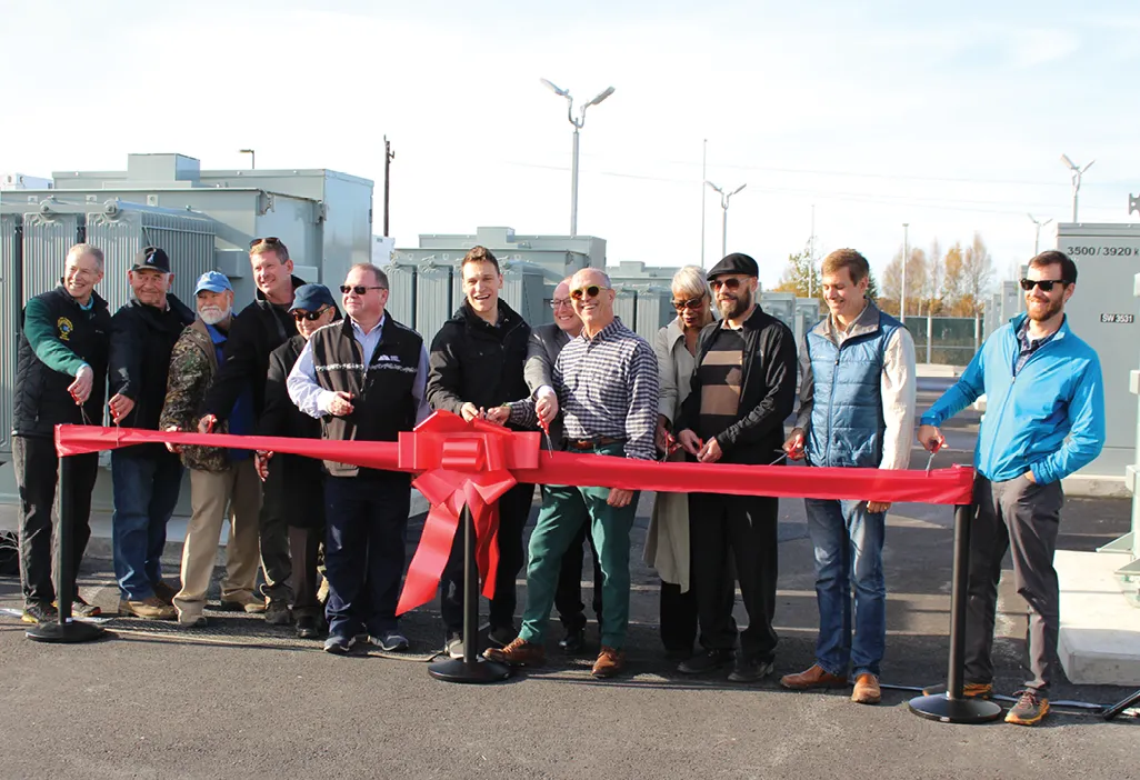 Group of people cutting a large red ribbon in front of utility equipment during a ceremony.