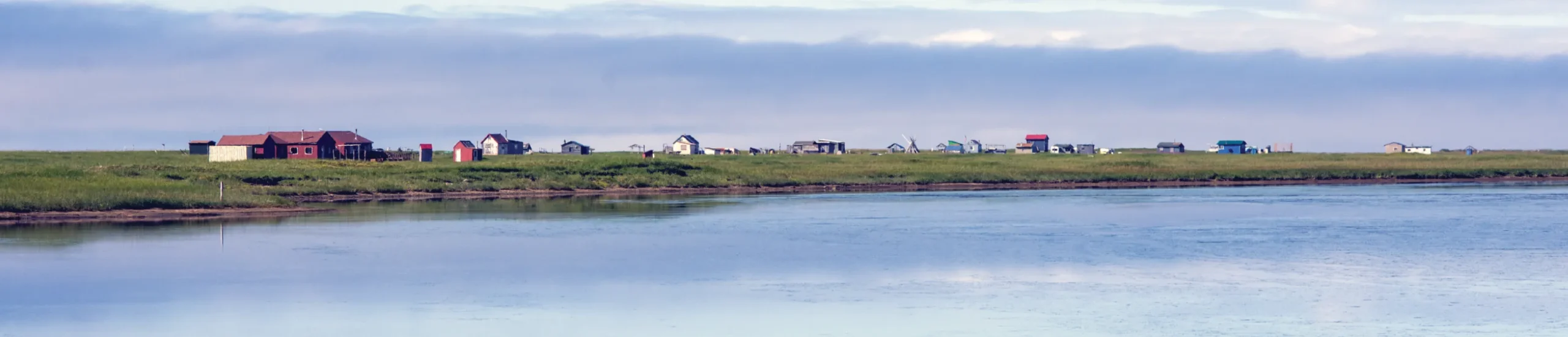 A calm river reflects a distant landscape with various buildings and green grass under a cloudy sky.