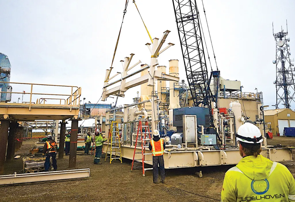 Construction workers operate a crane to lift equipment at an industrial site with machinery and ladders visible.