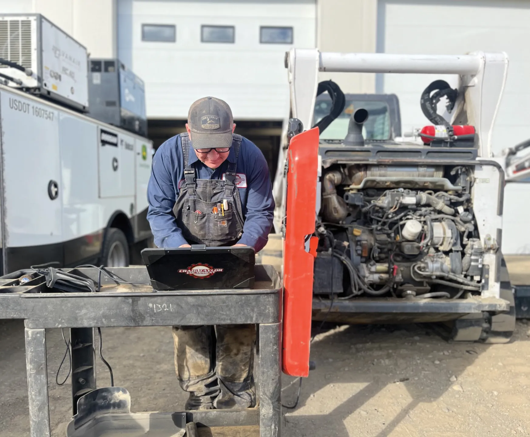A mechanic works on a laptop at a table next to an open engine compartment of a vehicle.