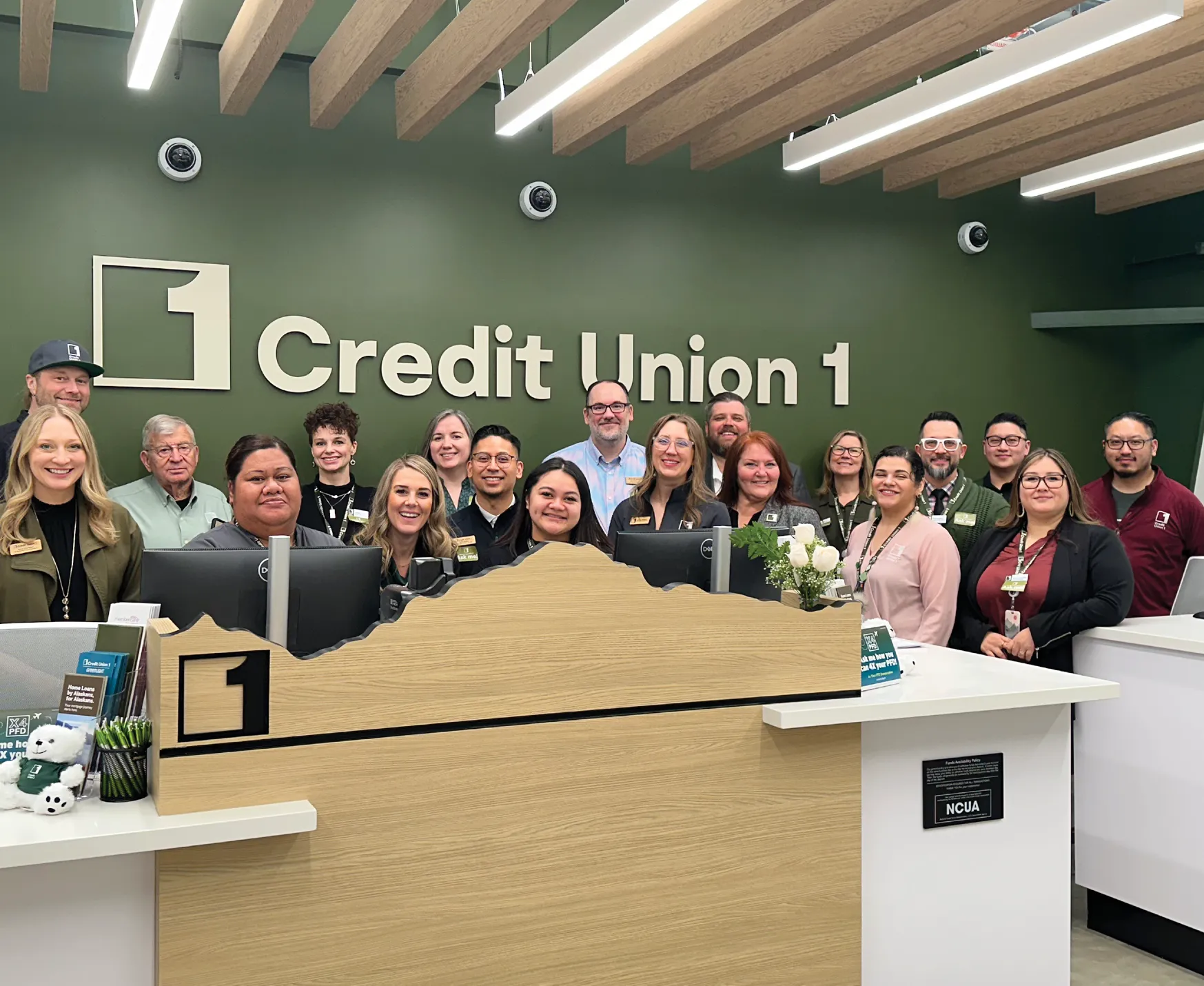 Group of employees posing together at Credit Union 1, with a green wall and reception desk in the background.