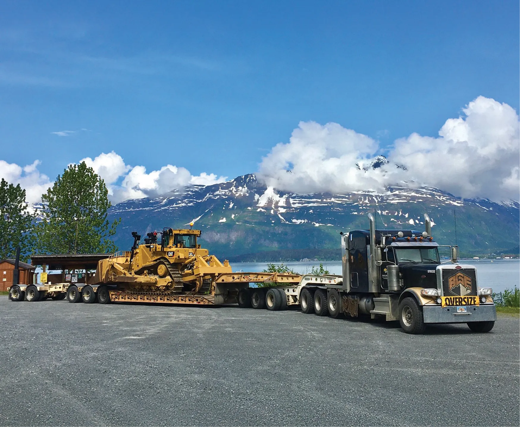 A large yellow bulldozer on a flatbed truck with an "OVERSIZE" sign, set against a mountainous landscape and blue sky.