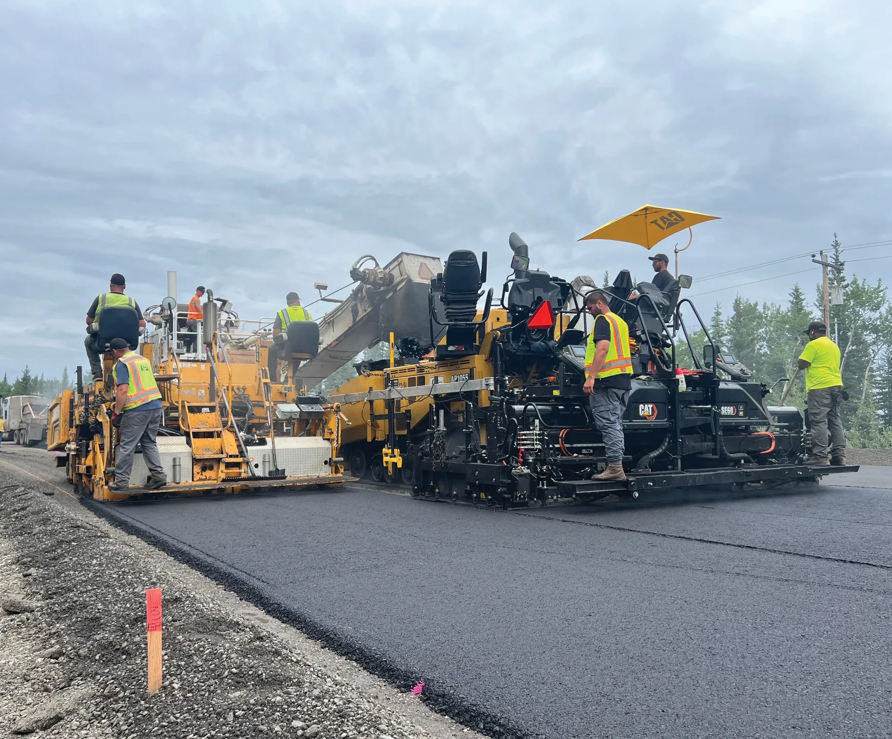 Construction workers operating heavy machinery to pave a road, with a cloudy sky in the background.