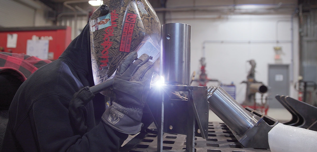 A welder in a dark hoodie and helmet with stickers works on a metal piece, bright sparks flying from the welding torch.