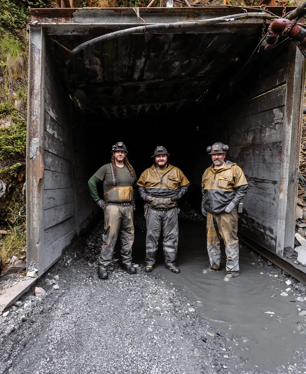 Three miners in work gear and helmets stand at the entrance of a dark mine tunnel, with one side of the tunnel floor covered in water and the other in gravel.