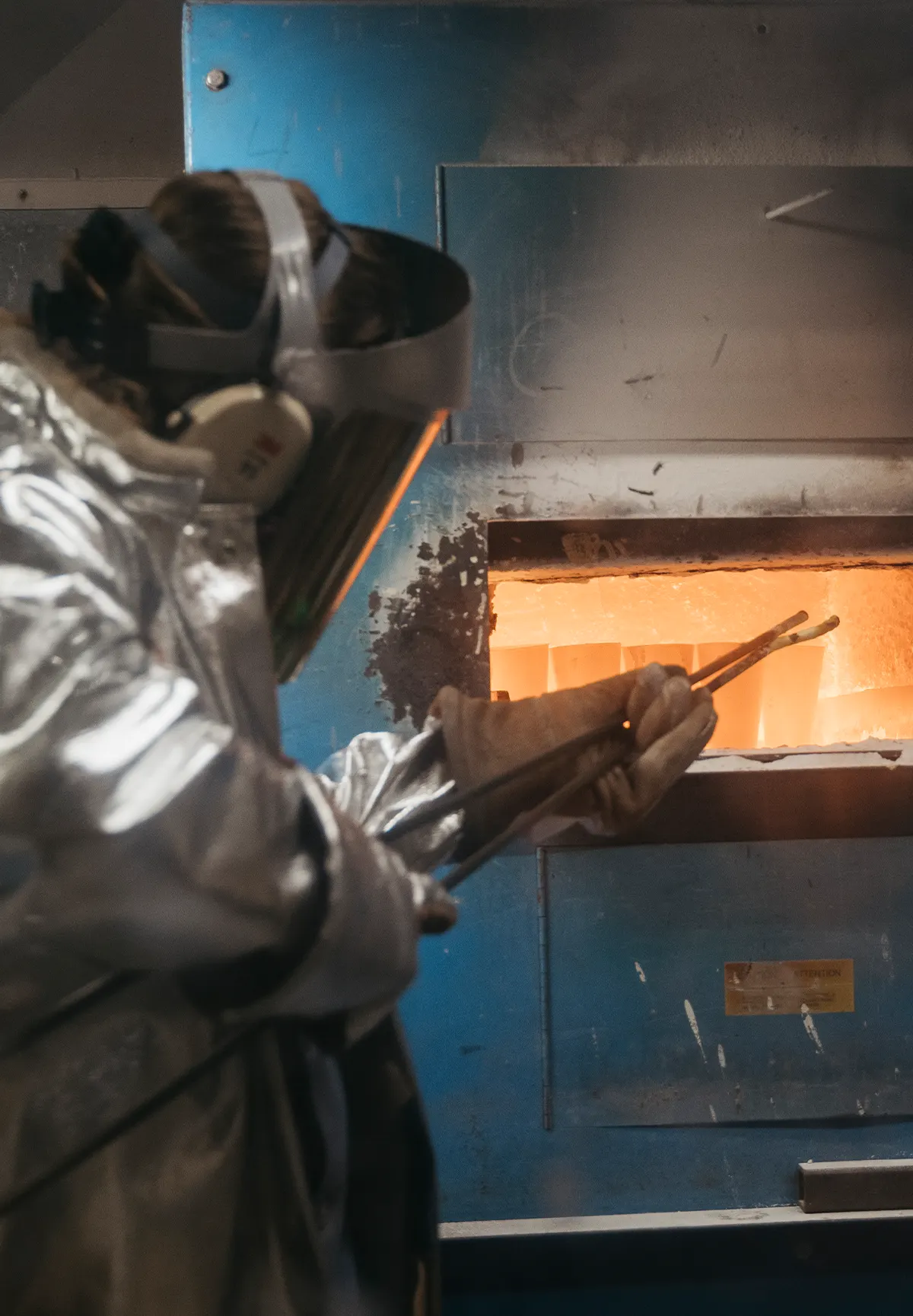 A person in a silver heat-resistant suit, face shield, and ear protection uses tongs to handle glowing material inside a high-temperature furnace, performing fire assay analysis to determine precious metal content.