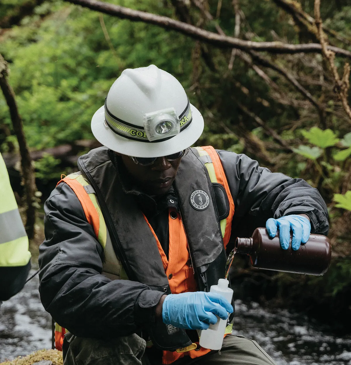 A person in a hard hat, safety vest, and gloves kneels by a stream, carefully pouring water from a brown bottle into a smaller white sample bottle, surrounded by lush greenery.