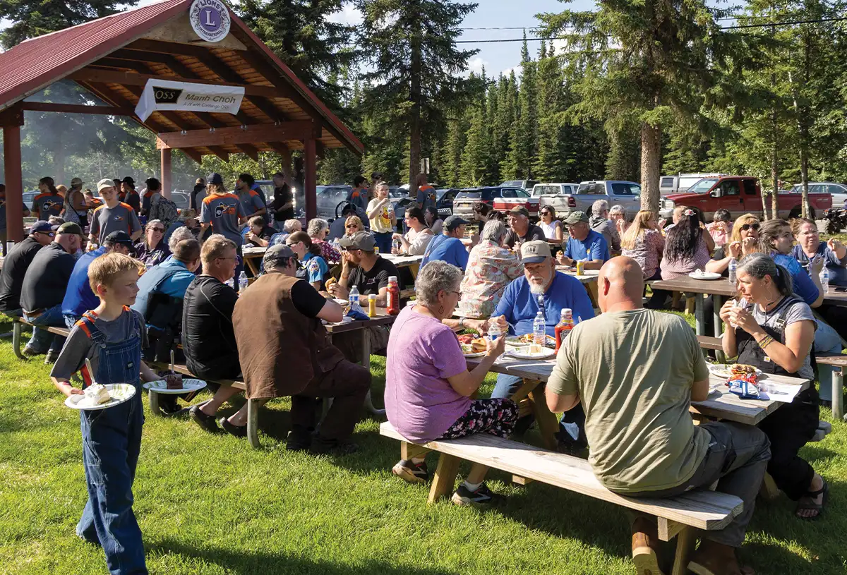 A sunny outdoor scene with many people gathered under a wooden pavilion and around picnic tables on a grassy field, enjoying a barbecue.
