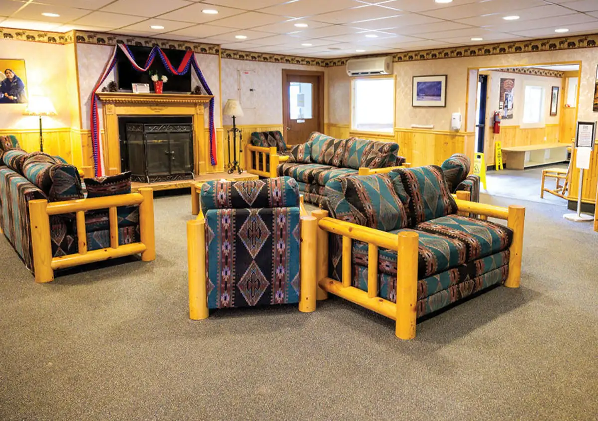 A cozy lodge-style living room with three patterned sofas, a wooden fireplace, and wood-paneled walls, featuring a warm and inviting atmosphere.