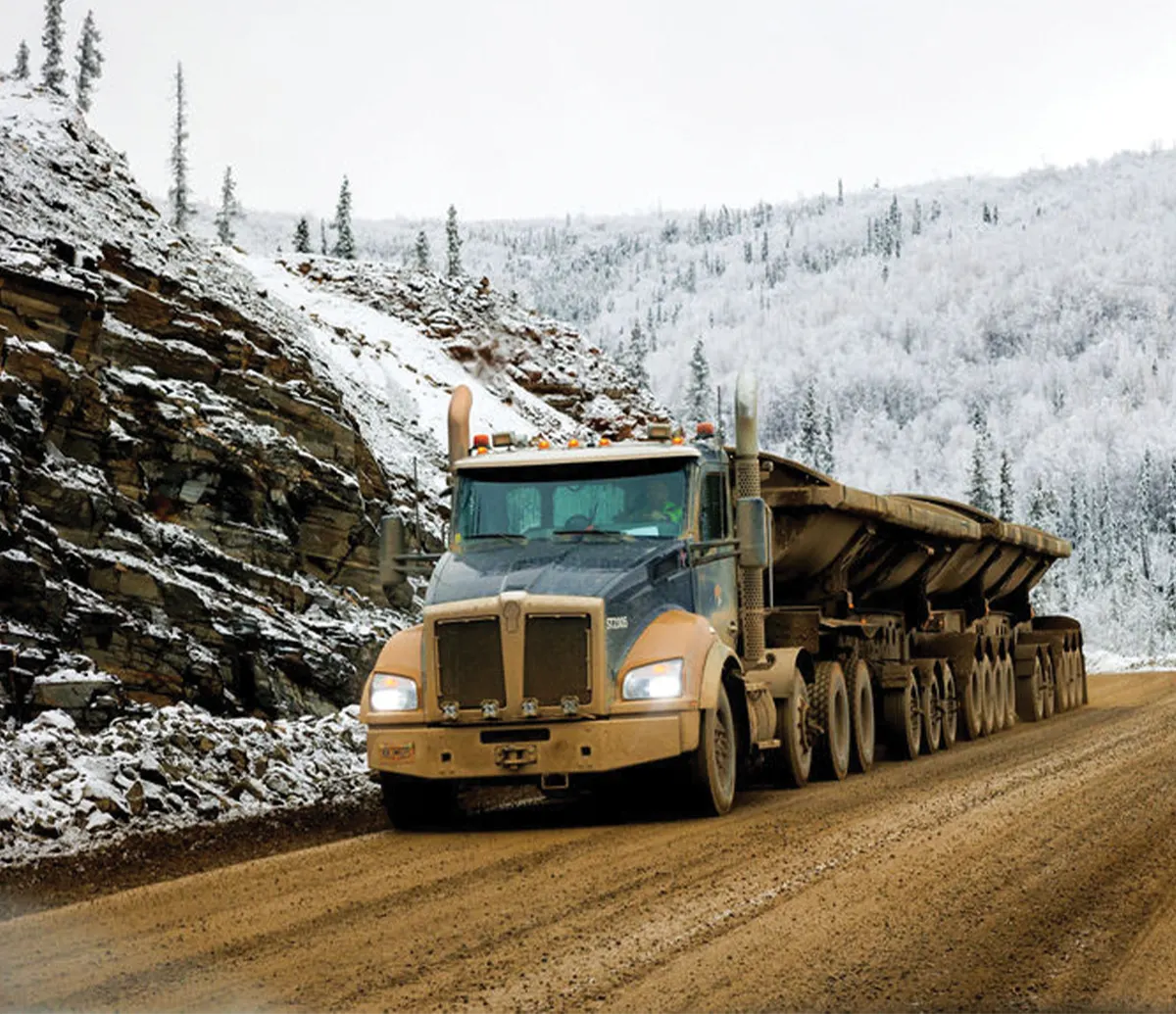 A large, heavy-duty haul truck with multiple axles and a dirty exterior drives along a muddy road, with a snow-covered rocky hillside on the left and a forest of snowy trees in the background.