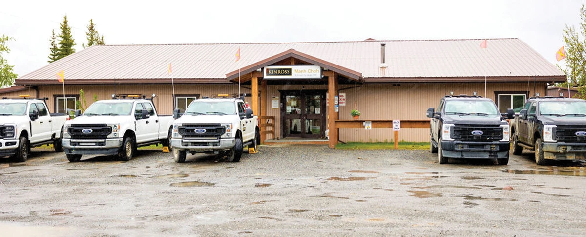 A row of white and dark pickup trucks parked in front of a long, single-story brown building with a central entrance under a small gable, displaying a “KINROSS Manh Choh” sign.
