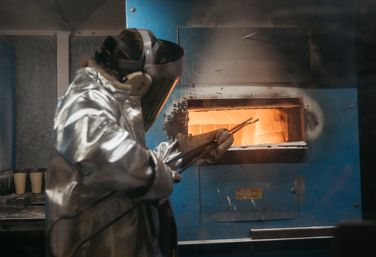 A person in a silver heat-resistant suit, face shield, and ear protection uses tongs to handle glowing material inside a high-temperature furnace, performing fire assay analysis to determine precious metal content.