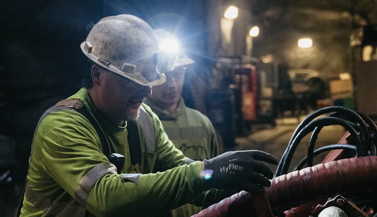 Two miners in hard hats with headlamps are visible in a dimly lit underground mine, with the foreground miner in a lime green shirt and gloves appearing to work on machinery.