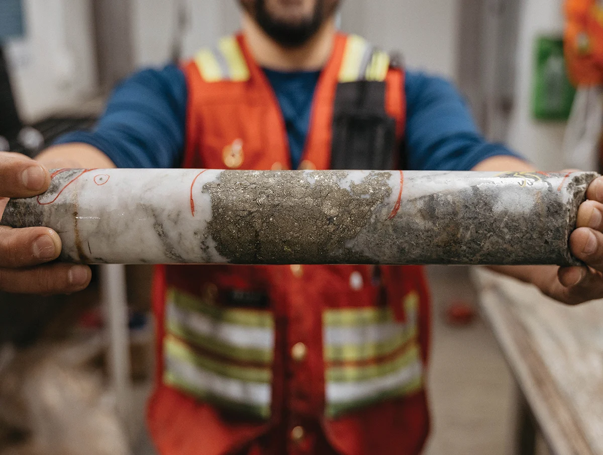 A person wearing a red safety vest holds a long, cylindrical core sample, which is half white rock and half dark, granular material, marked with red lines.