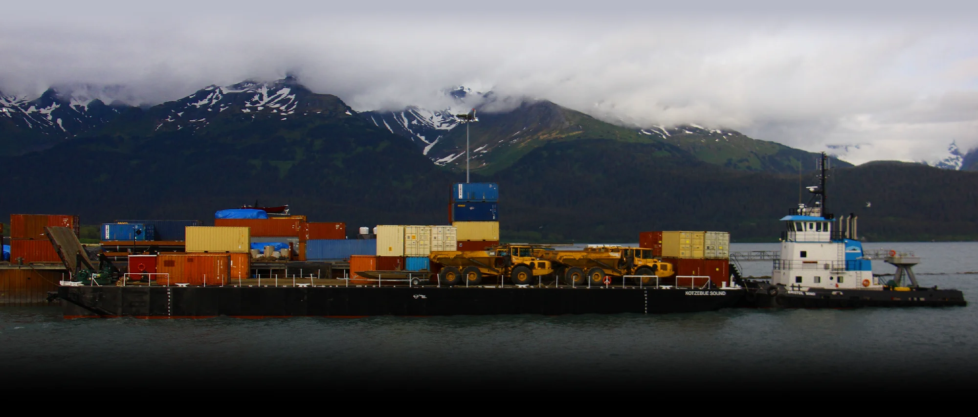 Tugboat pushing a cargo barge loaded with shipping containers and heavy yellow construction equipment in Cook Inlet, Alaska, with snow-capped mountains in the background