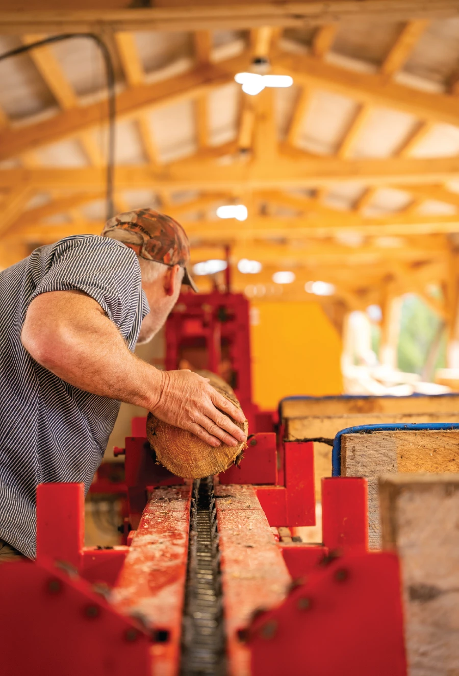 Man in a cap and striped shirt placing a small log onto a saw carriage inside a wooden-framed small-scale timber mill