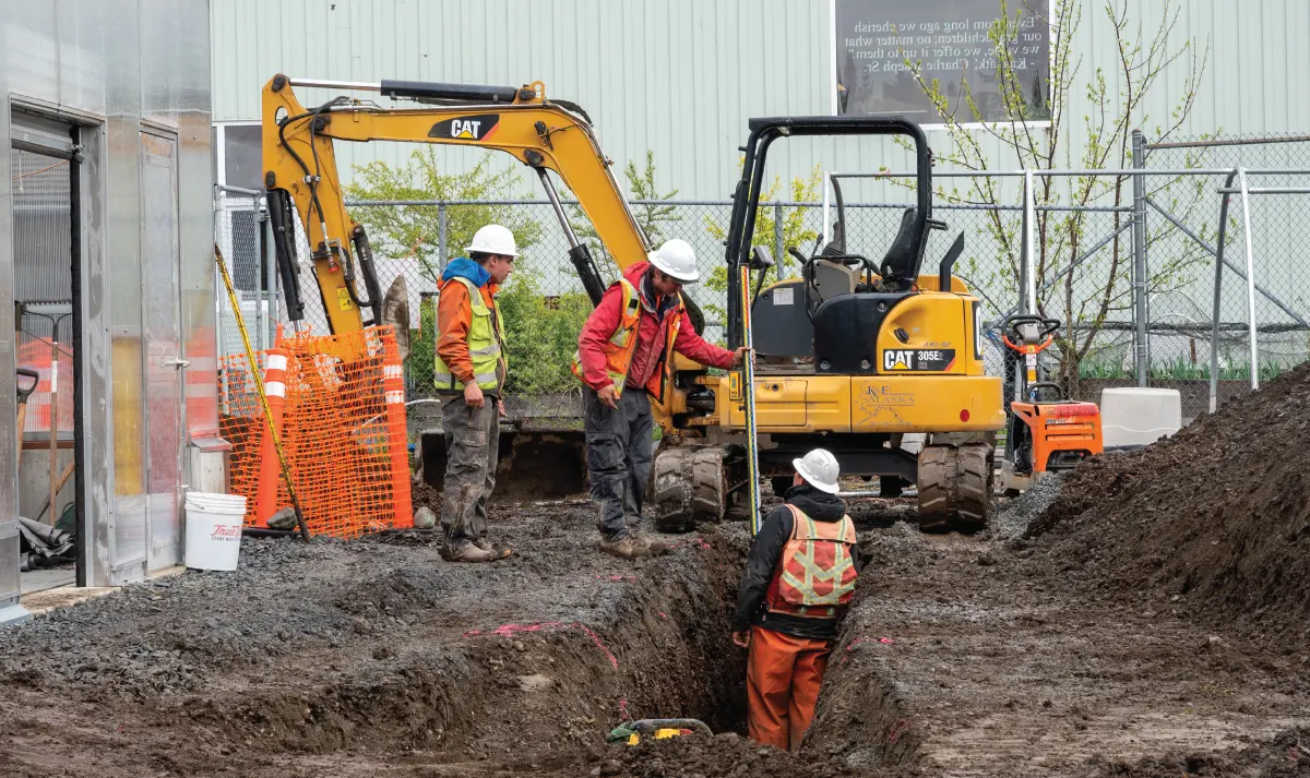 Construction crew using a CAT mini-excavator to dig a trench, with one worker taking measurements at the excavation site