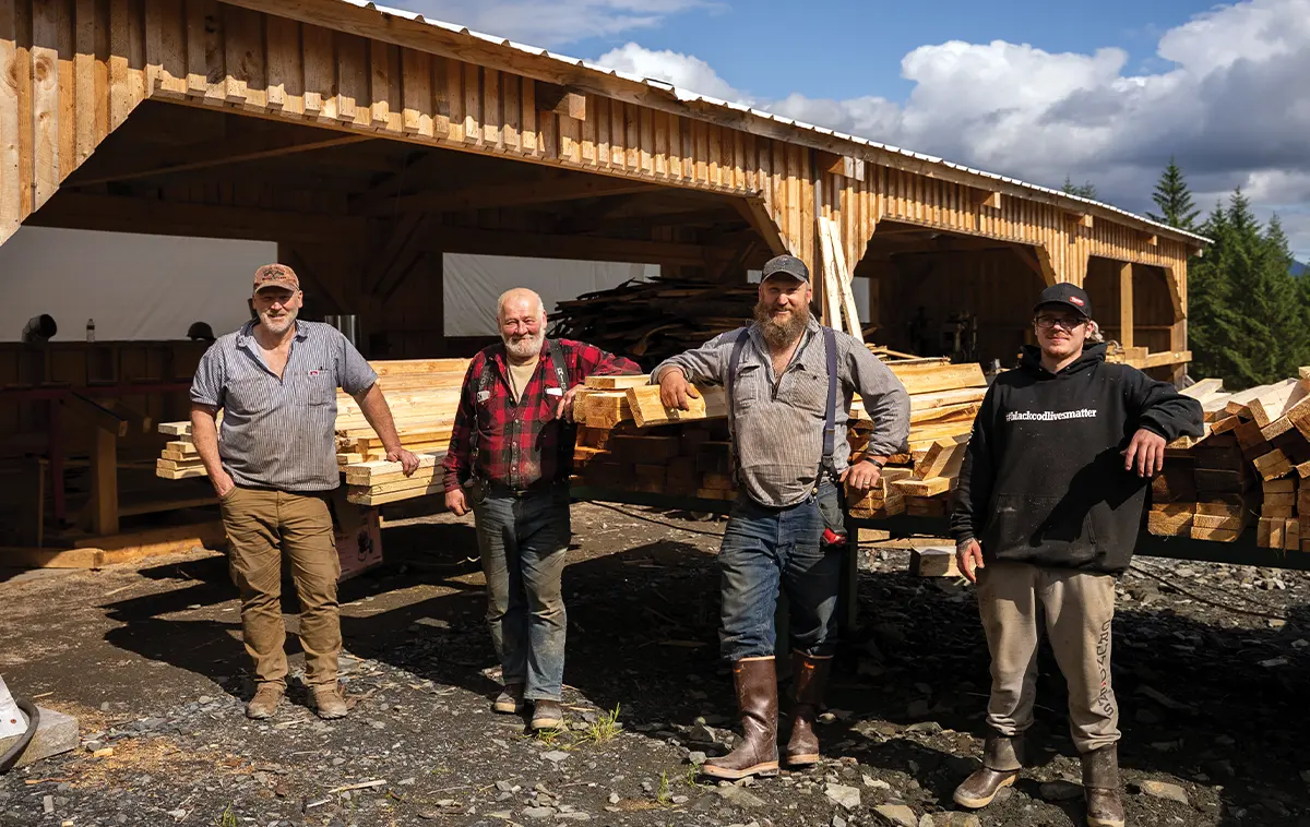 Four sawmill workers posing proudly in front of a timber structure and freshly cut wood stacks at their small-scale lumber operation