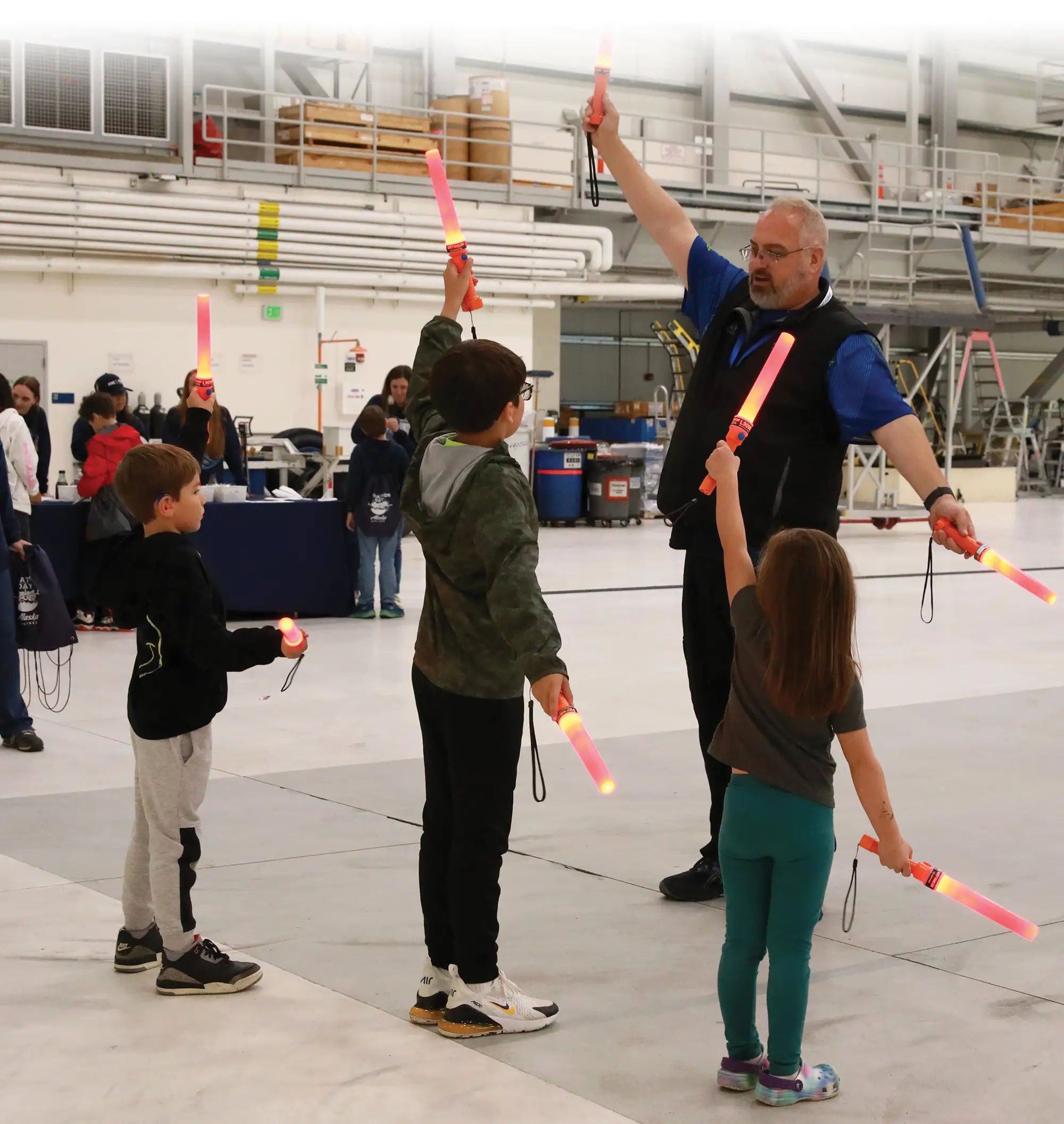 Adult instructor demonstrating airport marshalling signals with light wands to three children inside an aircraft hangar or maintenance facility