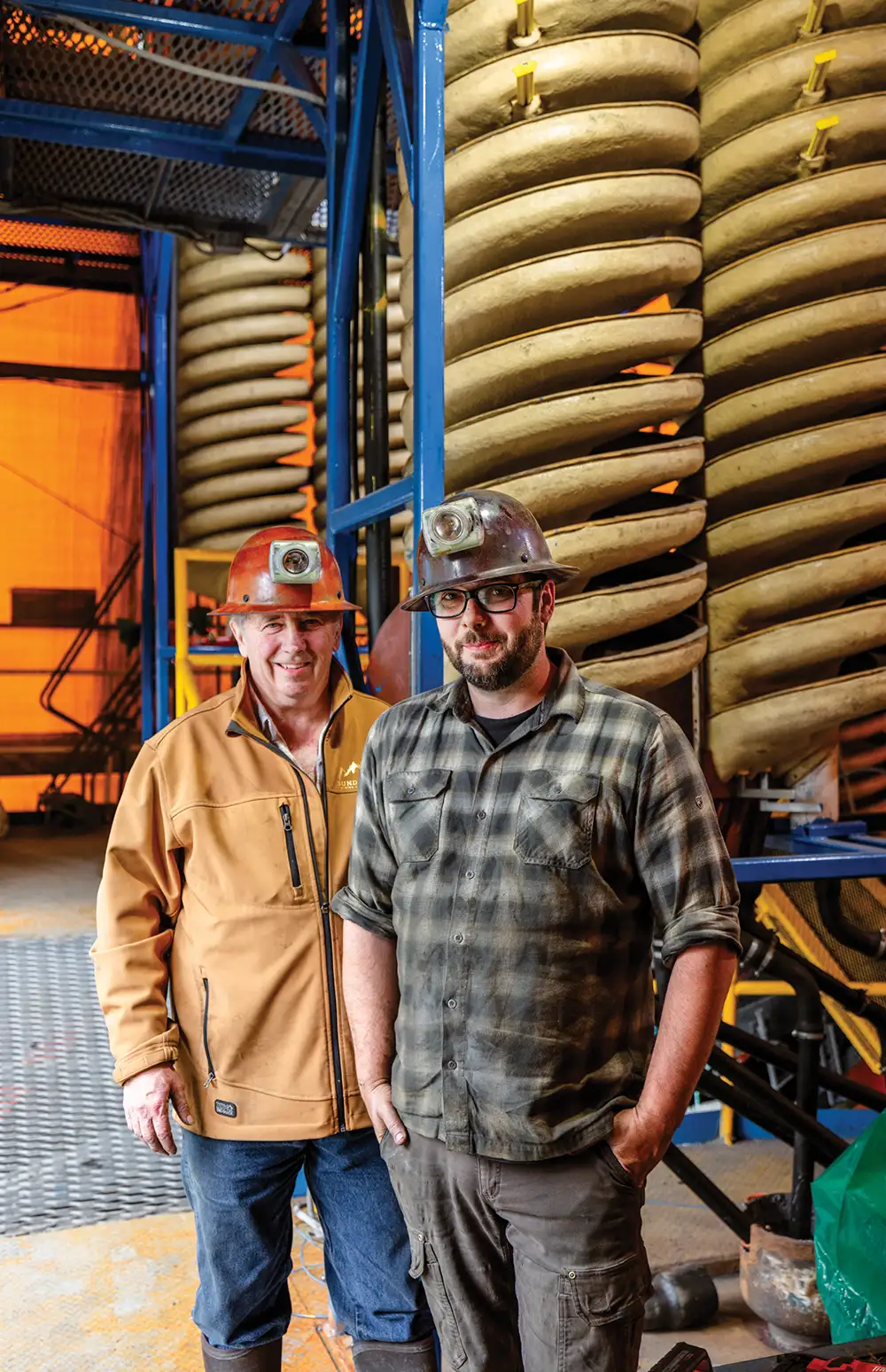 Two miners in hard hats stand in a processing facility in front of large spiral concentrators.