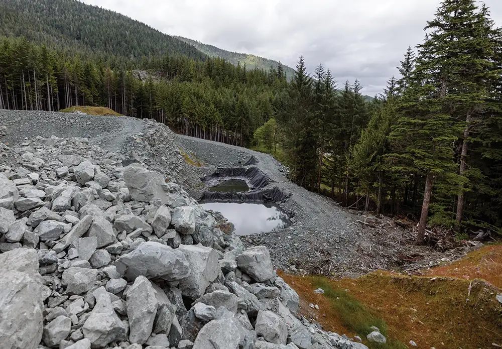 An aerial view of a forested hillside with a terraced, rocky mining area leading down to two small, lined retention ponds.