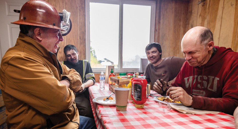 Four men, some wearing work attire and a hard hat, smile and laugh while eating a meal at a table with a red and white checkered tablecloth.