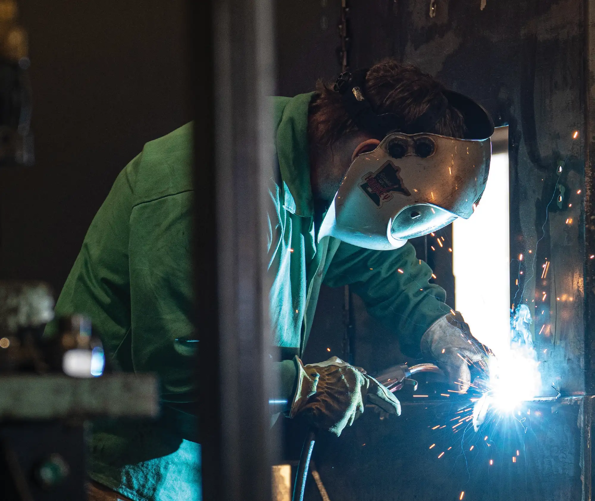 A welder wearing a green welding jacket and a protective helmet is shown in profile, concentrating on the bright arc light and sparks created while working on a metal piece in a dim industrial setting.