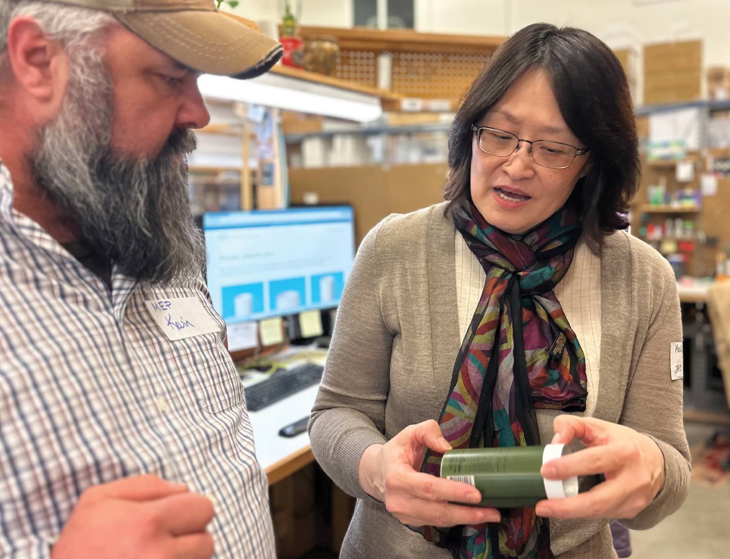 A close-up photograph shows two people in an office or workshop setting, focused on a small, olive-green cylindrical product that the woman is holding. The woman on the right, wearing a scarf and glasses, appears to be explaining the product to the man on the left, who has a long gray beard and is wearing a baseball cap and a plaid shirt with a name tag that reads "MEP Kevin."