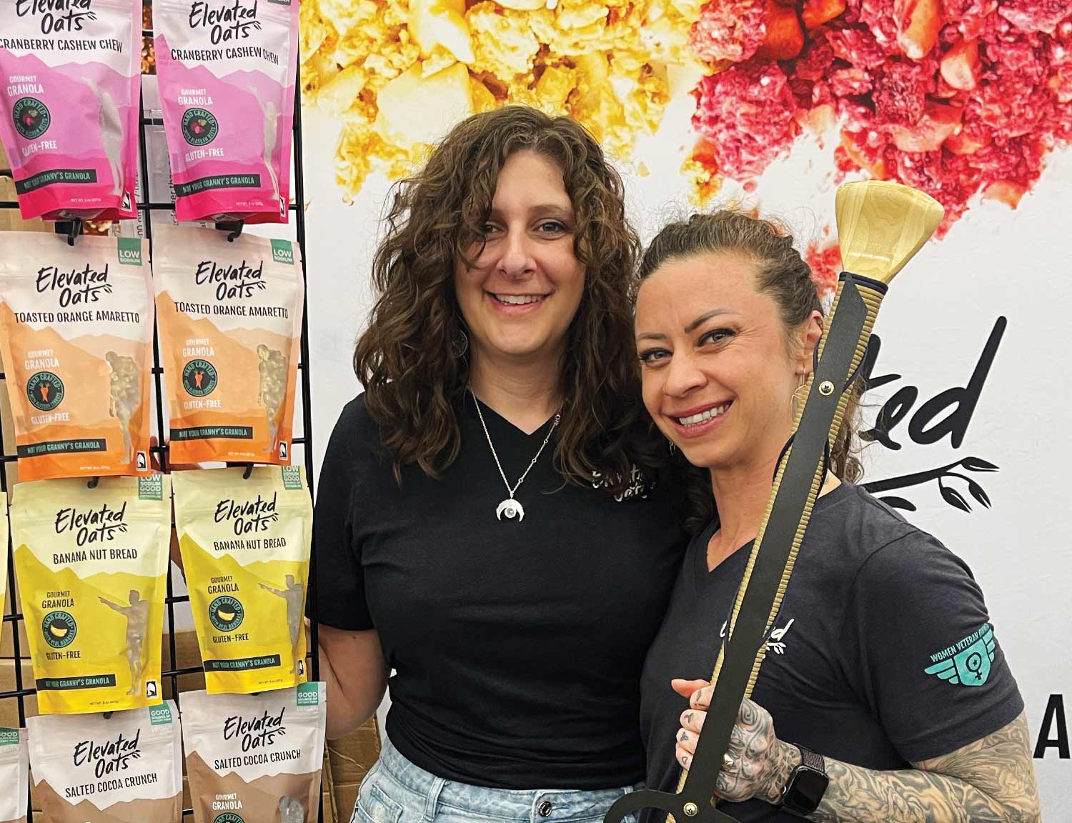 A photo features two smiling women standing in front of a shelf stocked with various flavors of "Elevated Oats" granola in brightly colored, stand-up pouches. The woman on the right has tattoos and is holding a decorative, wooden award.
