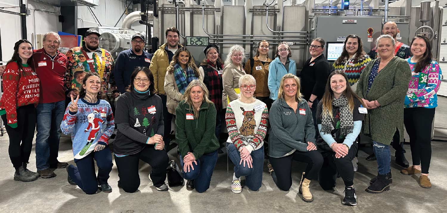 A diverse group of approximately 20 staff members pose for a group photo in what appears to be a food production or manufacturing facility, with industrial equipment visible in the background. Many of the staff, who are standing and kneeling, are wearing Christmas sweaters or other festive attire.