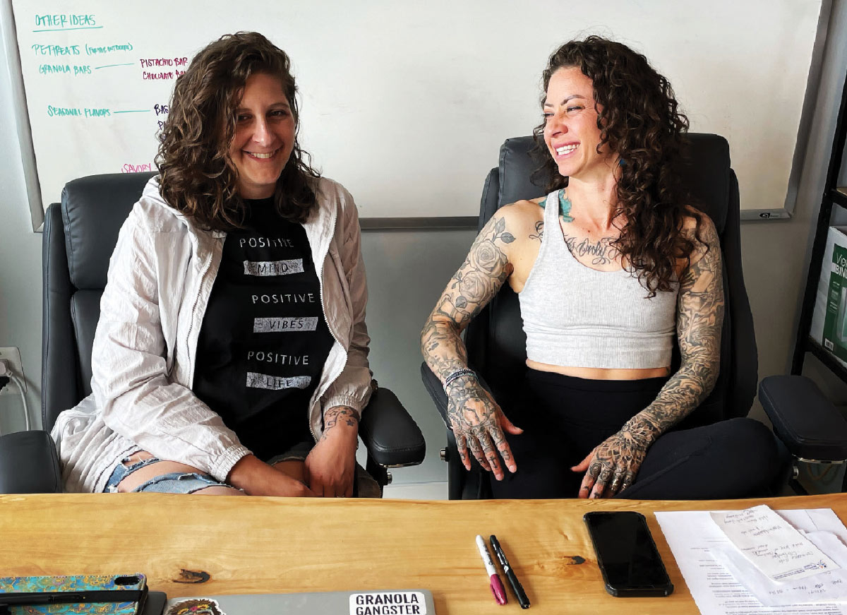 A photo features two women seated in office chairs behind a wooden desk, both smiling. The woman on the left is wearing a light jacket over a black t-shirt that reads "POSITIVE VIBES POSITIVE LIFE," while the woman on the right is wearing a light gray tank top. A whiteboard listing "OTHER IDEAS" is visible in the background.