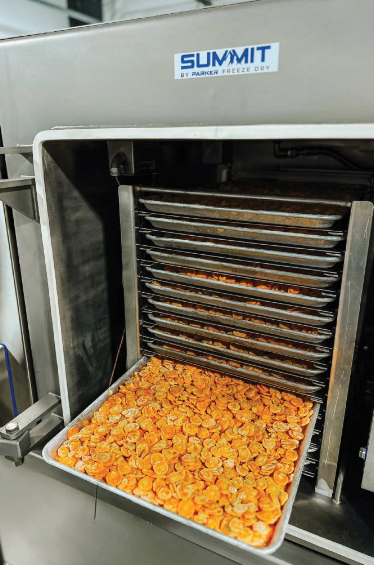 A tray filled with a single layer of freeze fried food is pulled out of a large, stainless steel "SUMMIT by PARKER FREEZE DRY" industrial machine. Multiple trays are stacked inside the unit above the exposed tray, showcasing the commercial-scale food processing equipment.