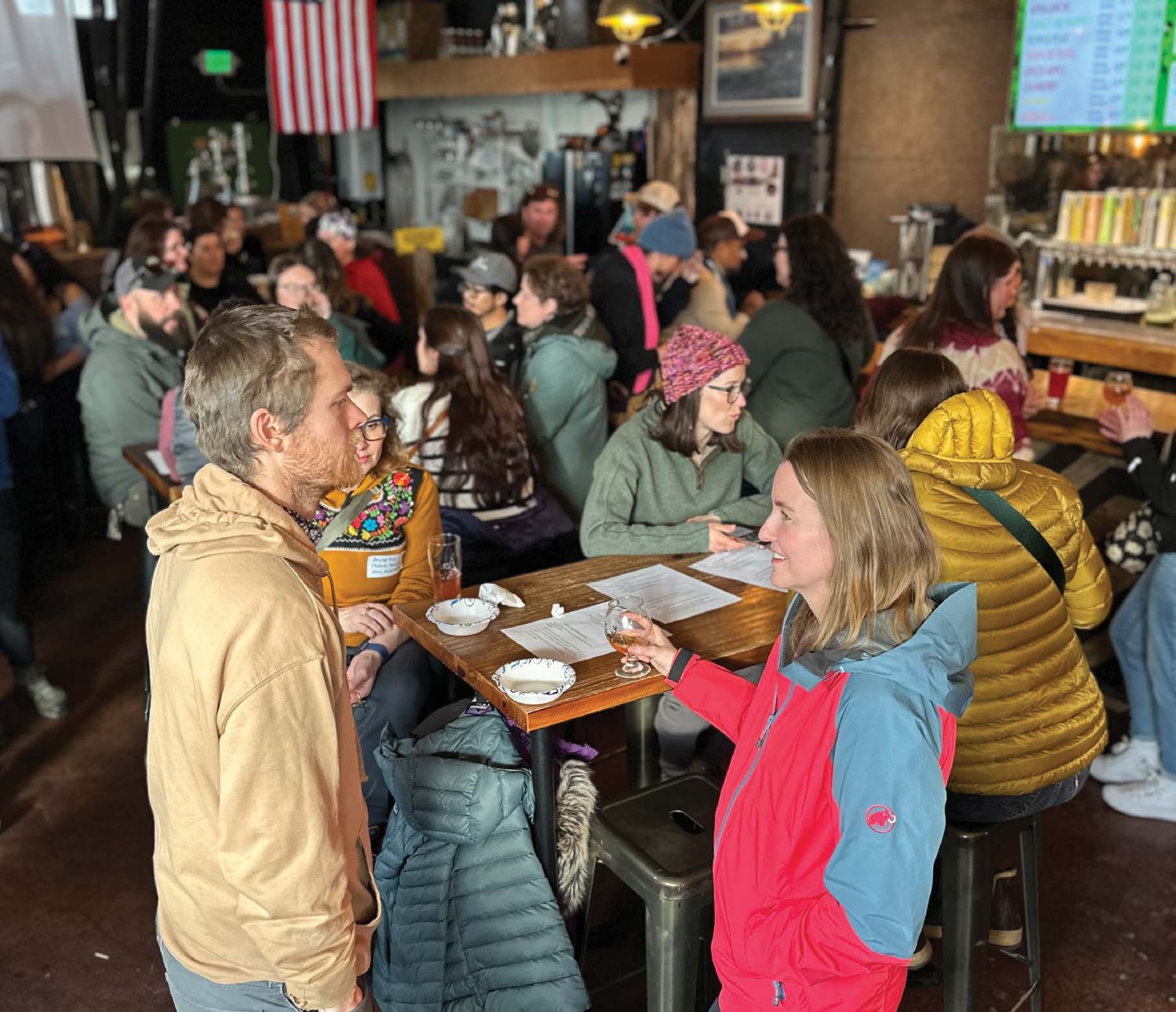 A photograph taken inside a dimly lit crowded brewery or pub shows two people in the foreground talking near a table with paperwork and drinks. The woman is smiling, wearing a bright red and blue jacket, and holding up a small glass of beer toward the man in a tan hooded sweatshirt, while many other patrons are visible seated at tables and the bar in the background.