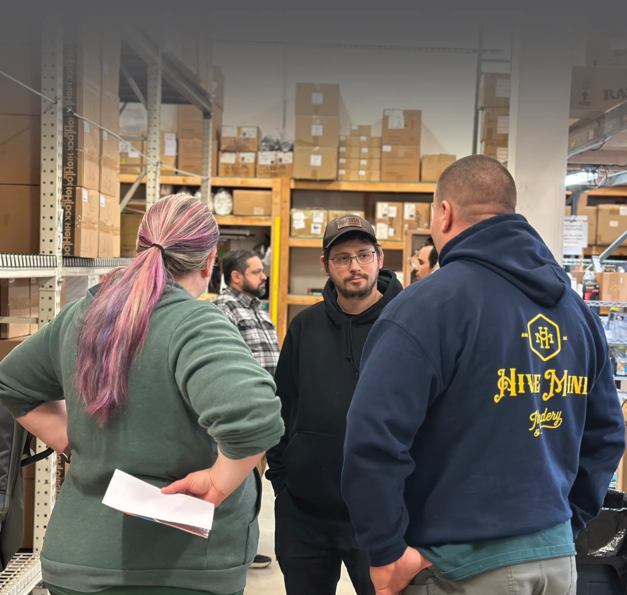 A photograph taken inside a warehouse or storage room shows a group of people having a discussion, surrounded by tall shelves stacked with cardboard boxes and inventory. In the foreground, a woman with purple-dyed hair faces a man in a black hoodie and a baseball cap, who is intently listening to a man in a dark blue "Hive Mind" hoodie seen from the back.