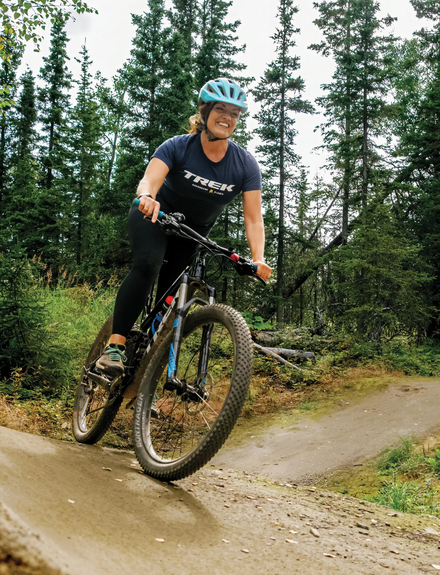 Portrait orientation outdoor photograph cropped view of Michele Parkhurst, a smiling woman wearing a baby blue colored mountain bike helmet and a dark navy blue colored graphic t-shirt with black yoga legging sweatpants plus dark grey colored fitness shoes as she is riding a mountain bike downhill on a dirt trail; The trail has a banked curve in the foreground and she is surrounded by a dense green forest of pine trees