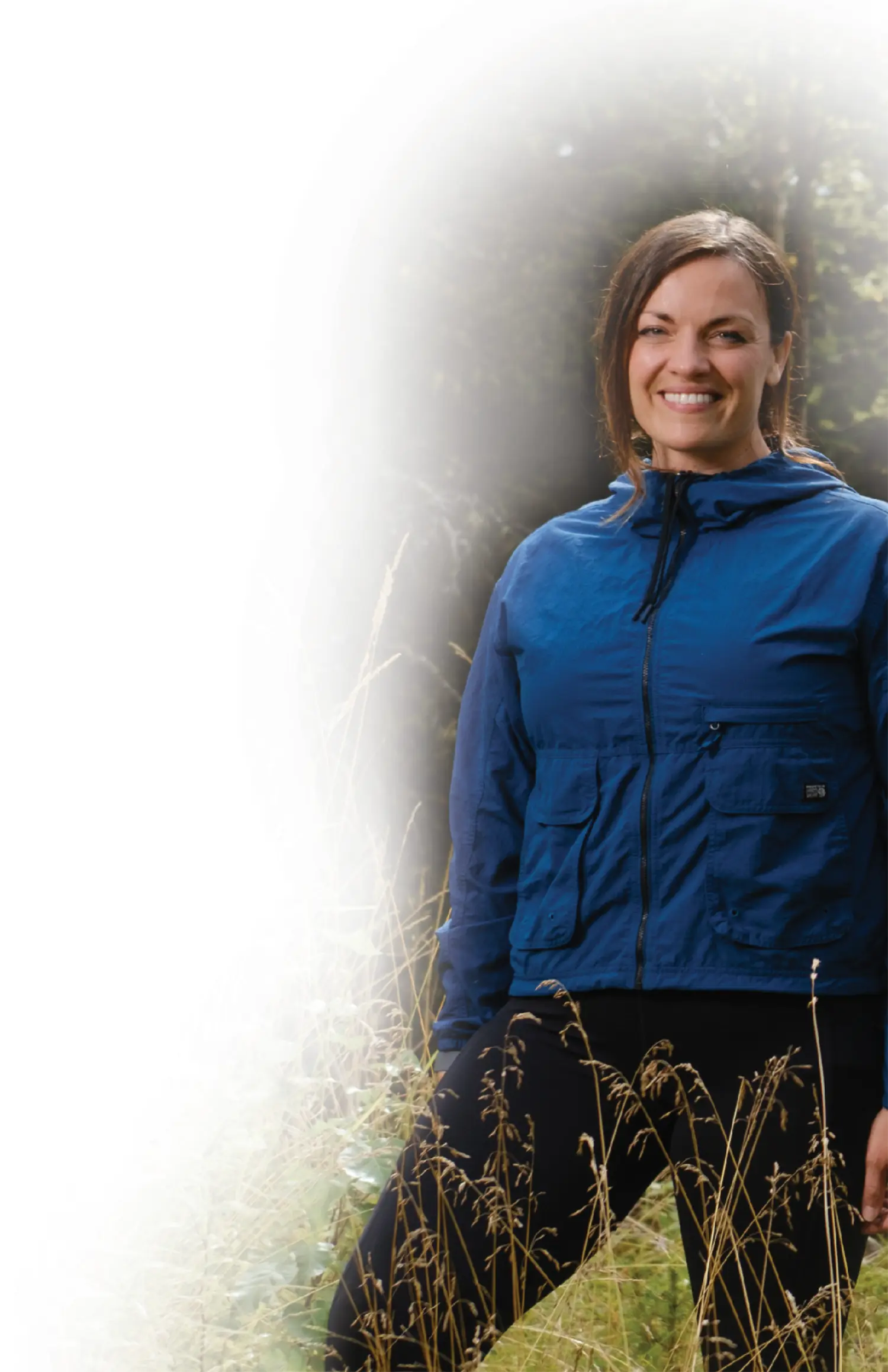 Outdoor photograph portrait orientation cropped view of Michele Parkhurst, a smiling woman with dark brown colored hair stands outside in a field of tall grass; She is wearing a dark blue colored thick jacket and black yoga legging sweatpants; The left side of the image is a fade-to-white digital gradient filter effect