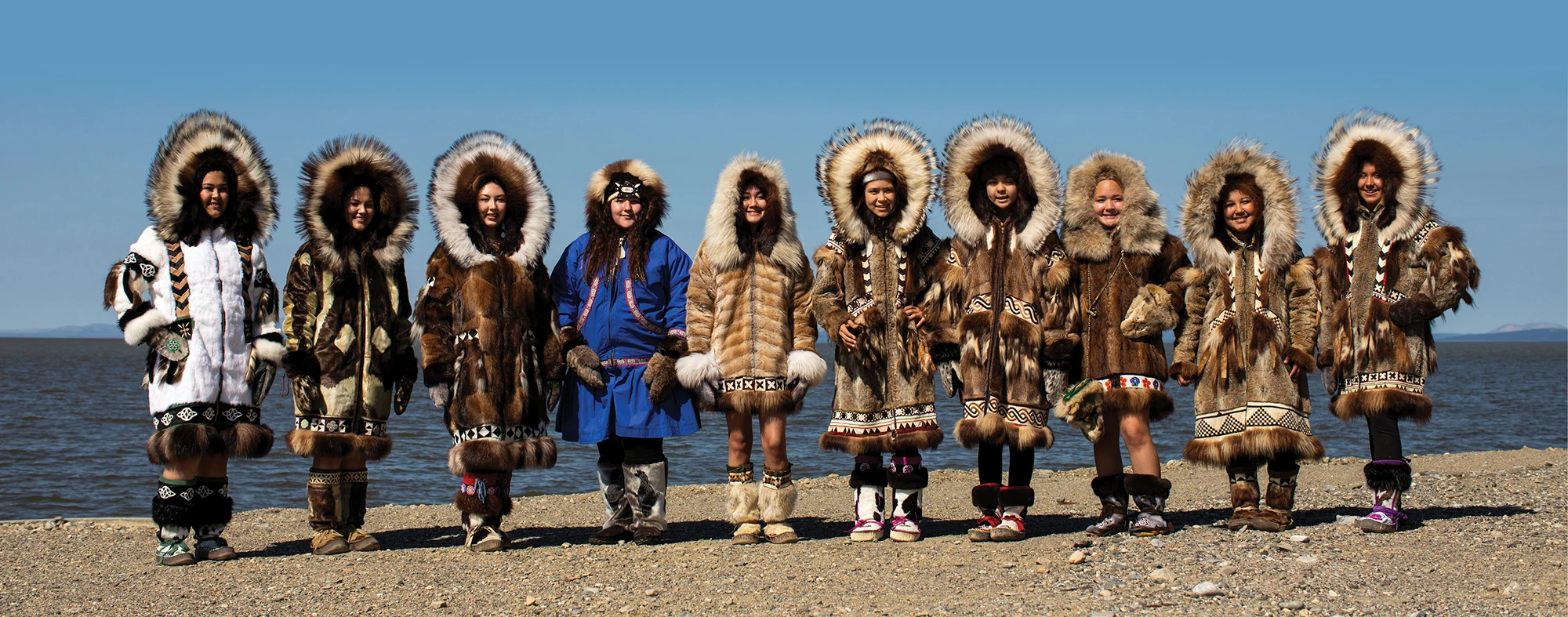 A group of nine people stands side by side outdoors by the shoreline, wearing traditional Alaska Native fur parkas with intricate patterns and large, fur-lined hoods. The sky is clear and blue, and the scene highlights cultural heritage and craftsmanship.
