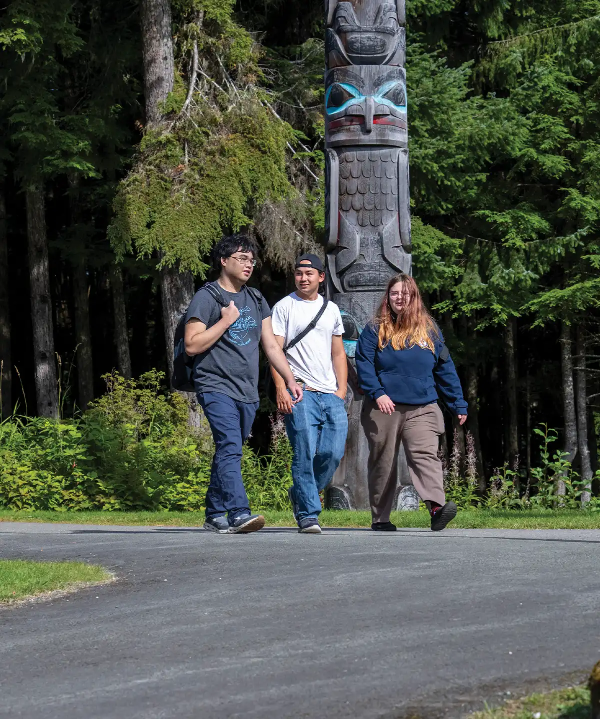 Three college aged young adults, two men and one woman, wearing backpacks walk down a paved path in a park with a large totem pole and tall trees in the background.
