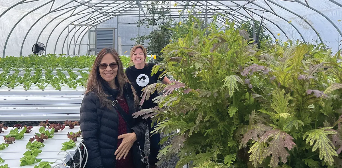 Two women stand inside a large, arched, plastic-covered hoop house (high tunnel) at Sitkalidak Sunrise Farm, surrounded by hydroponic lettuce on white racks and tall leafy green plants in the foreground.