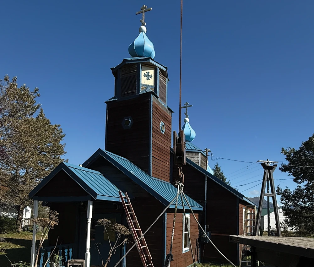 The historic Russian Orthodox church in Old Harbor, featuring dark brown wood siding and a steep blue metal roof, stands under a clear blue sky. It has two iconic blue onion domes topped with crosses.