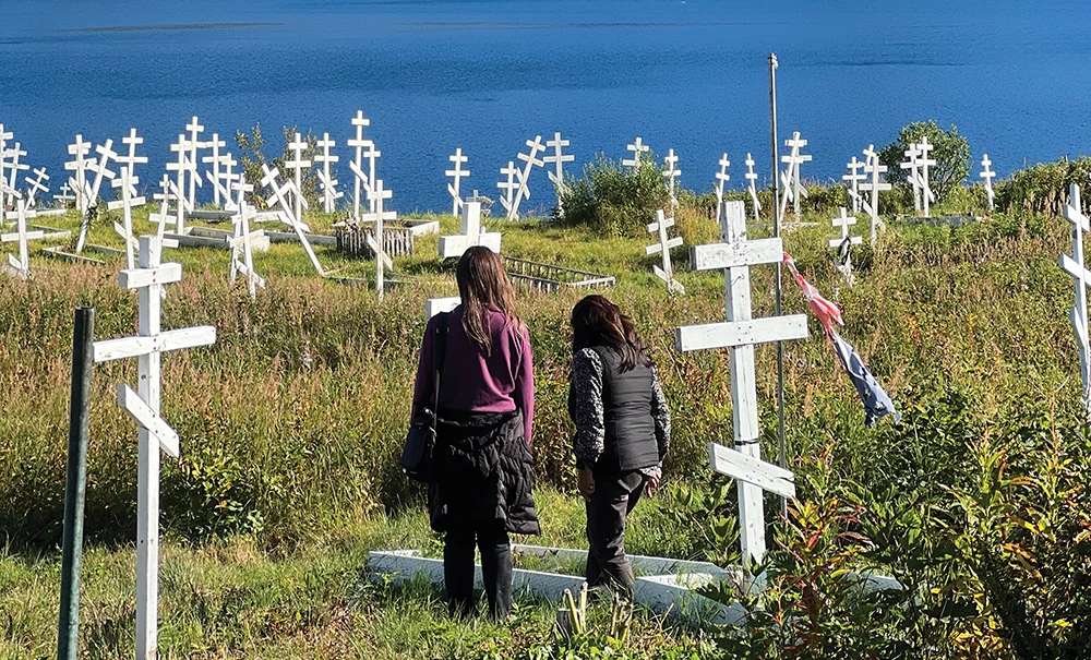 Two women stand with their backs to the camera, walking through a grassy cemetery on a hillside. Dozens of rustic white wooden Russian Orthodox crosses mark graves, with a large blue body of water in the background.