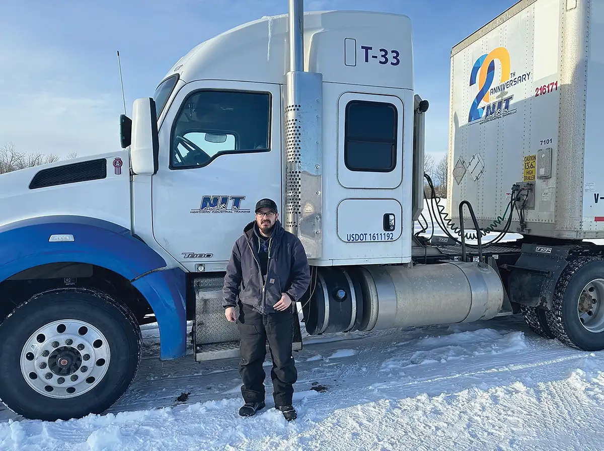 Northern Industrial Training (NIT) student or truck driver standing in front of a white semi-truck and trailer on a snowy road.