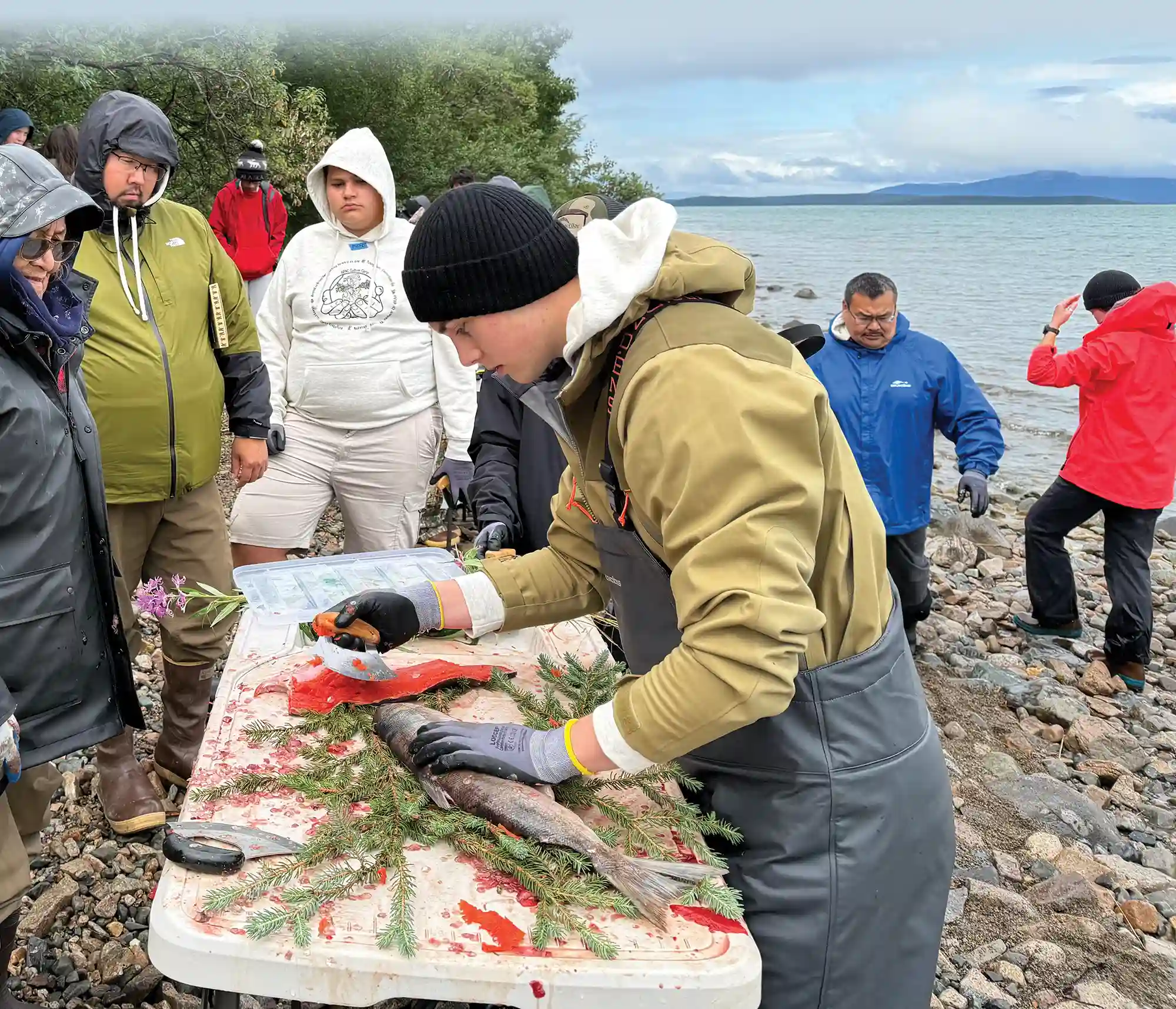 Young person wearing waders and gloves demonstrating the traditional technique of filleting a fish to a group of onlookers on a rocky beach.