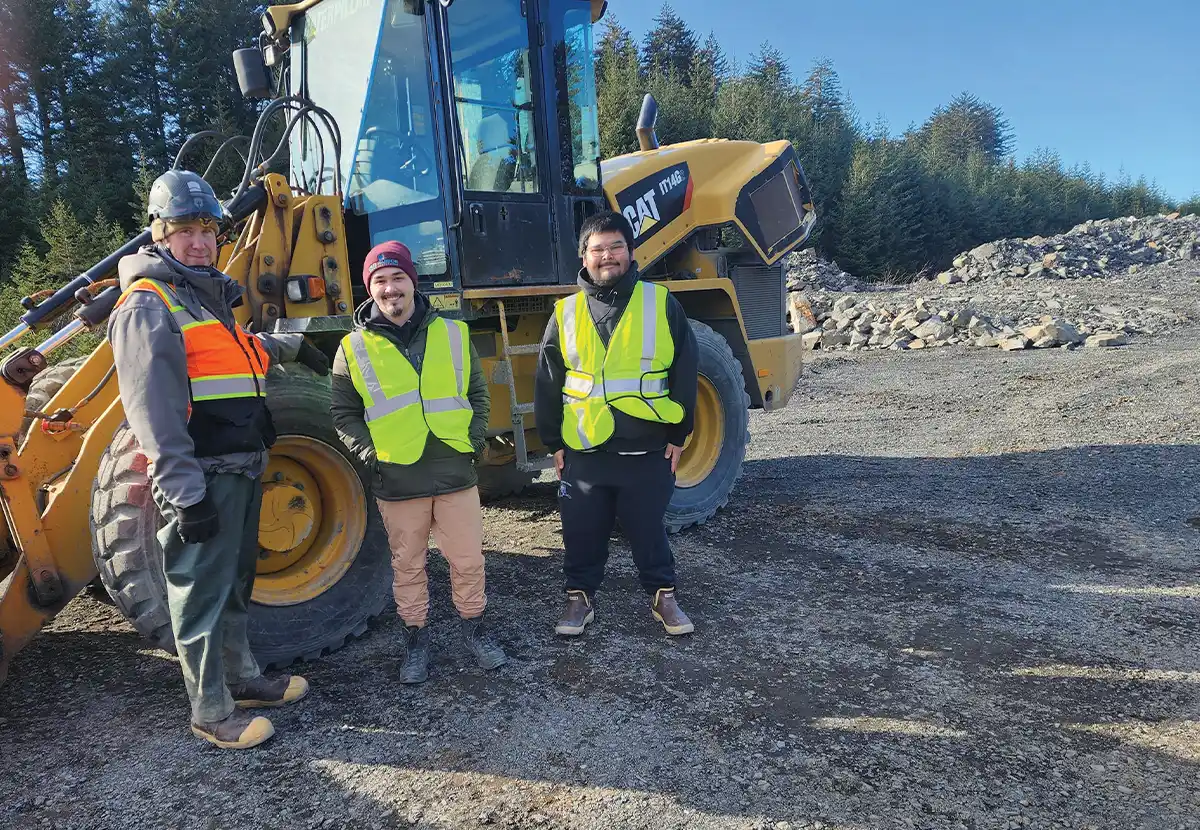 Construction crew of three men in safety vests standing next to a heavy equipment loader in a gravel pit.