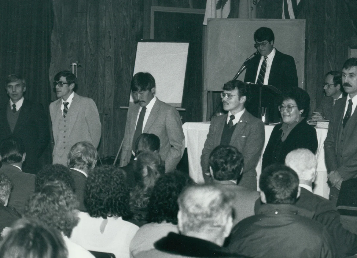 Historic photo of a business or community meeting with a panel of speakers and attendees, showing a man speaking at a podium